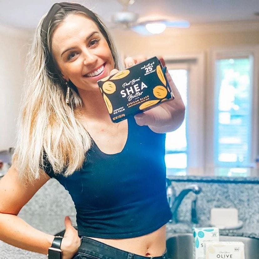 foreground woman with medium light complexion with blonde hair, a black fabric headband, cropped t-shirt and black jeans in a sunny, modern home standing in front of a granite countertop with a stainless steel sink holding a package of shea butter african black soap